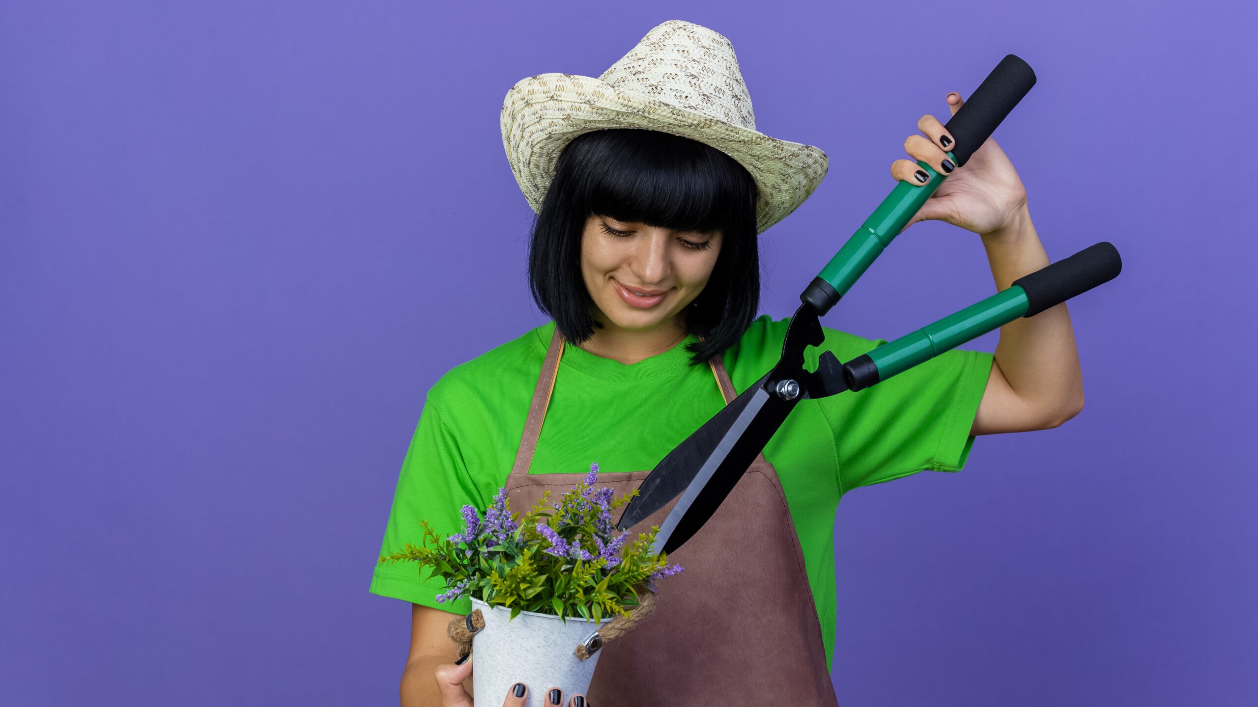 pleased young female gardener in uniform wearing gardening hat holds clippers over flowers in flowerpot isolated on purple background with copy space